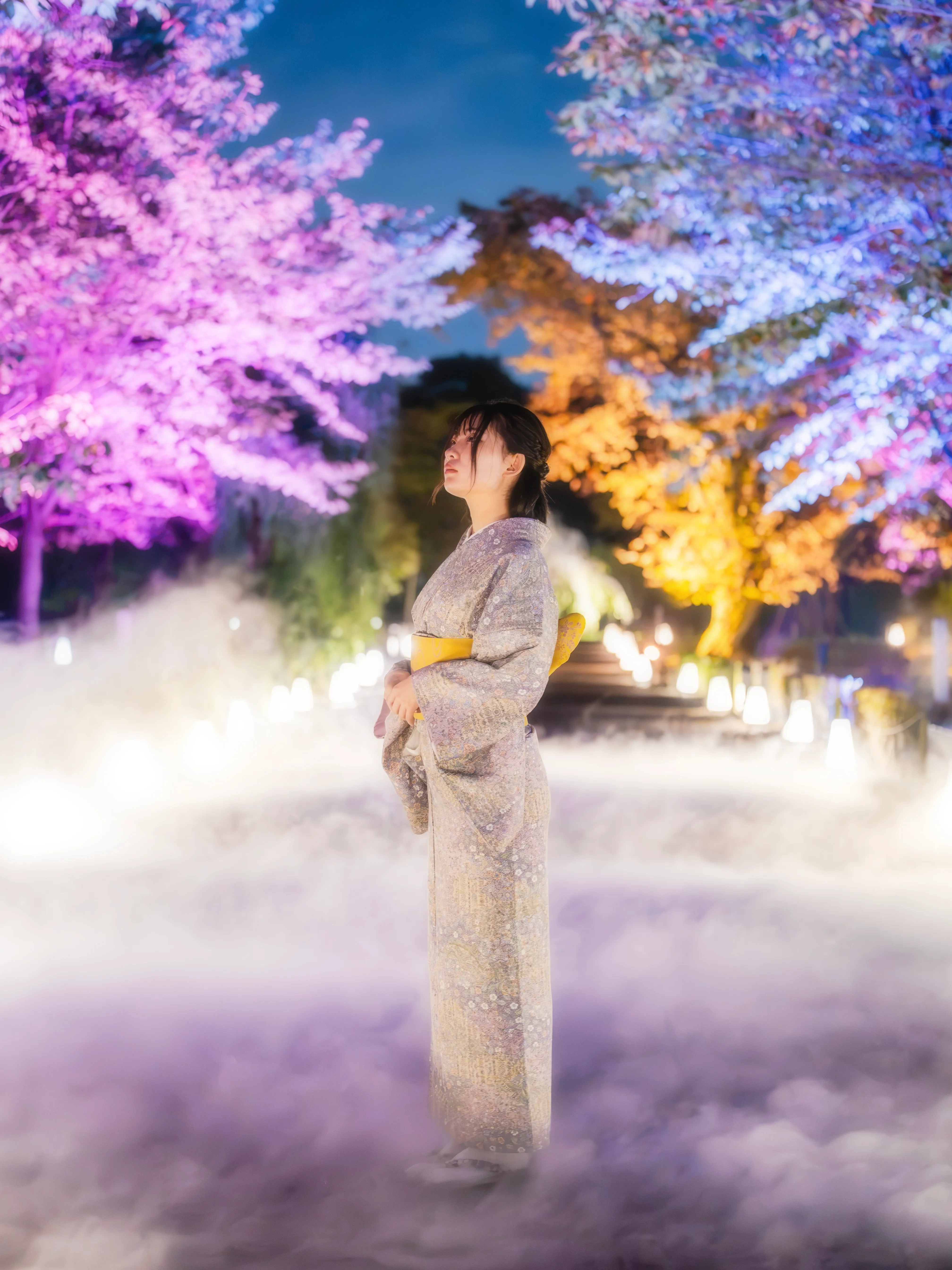 桃色と青に染まる木々を背に、霧の中に立つ着物姿の女性